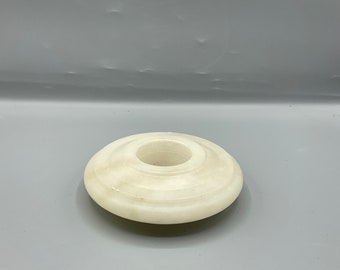 May include: A white, circular candle holder with a central hole. The candle holder is made of a smooth, light-colored material, possibly stone or marble. The design is simple and elegant, with a slightly raised outer edge.
