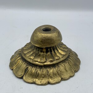 May include: A gold-colored, ornate base with a fluted, petal-like design. The base has a rounded top with a central hole. The metal has a slightly aged appearance.