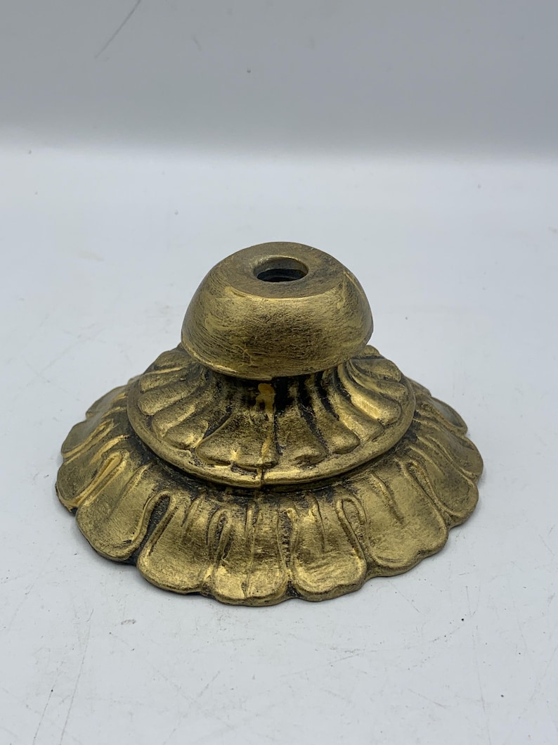 May include: A gold-colored, ornate base with a fluted, petal-like design. The base has a rounded top with a central hole. The metal has a slightly aged appearance.