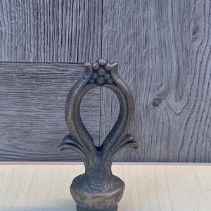 May include: A decorative metal finial with an ornate design. The finial is a dark bronze color and has a bulbous base.