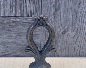 May include: A decorative metal finial with an ornate design. The finial is a dark bronze color and has a bulbous base.