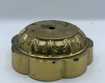 May include: A brass-colored, decorative base with a floral design around the top edge. The base has a scalloped edge and a hole in the center. The metal shows signs of wear and age.