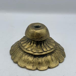 May include: A decorative, antique-style lamp base in a metallic gold finish. The base features a layered design with a fluted, petal-like lower section and a smaller, rounded top with a central hole. The base is designed to support a lamp.