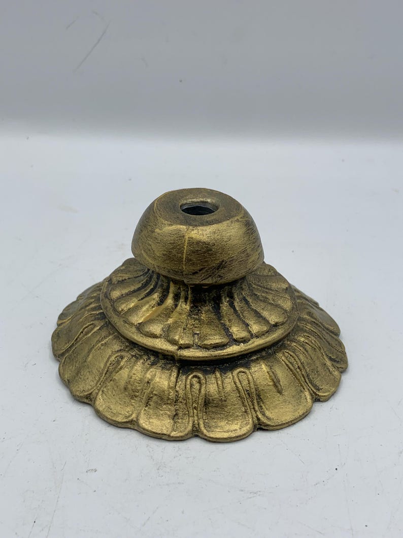 May include: A decorative, antique-style lamp base in a metallic gold finish. The base features a layered design with a fluted, petal-like lower section and a smaller, rounded top with a central hole. The base is designed to support a lamp.
