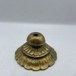 May include: A gold-colored, decorative metal stand with a tiered, floral-like base and a small, rounded top. The stand has a distressed finish, giving it a vintage appearance. The base has a series of detailed, petal-shaped edges.