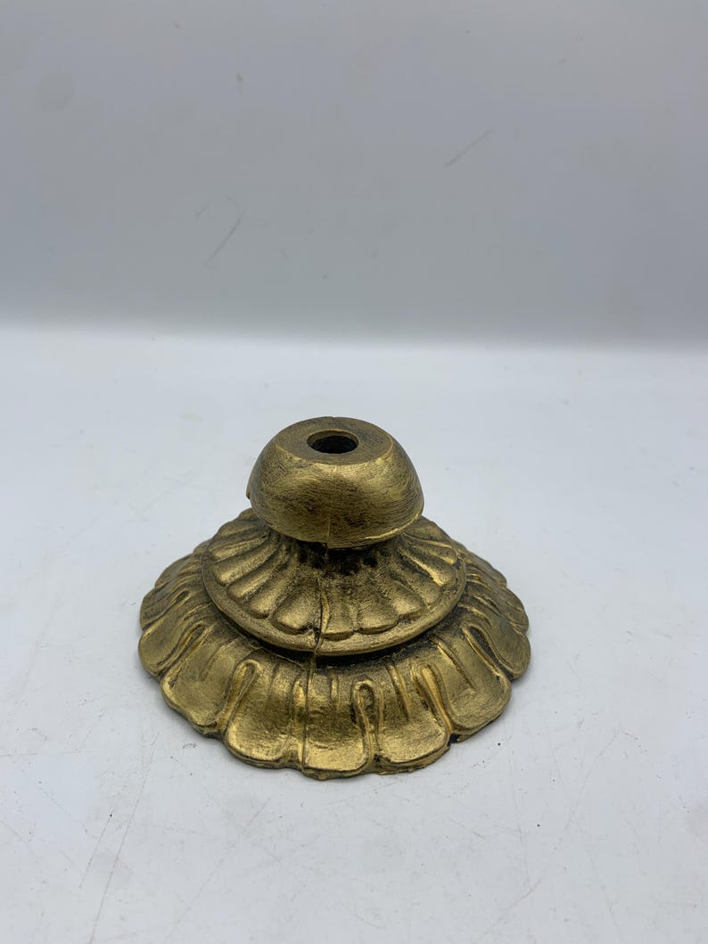 May include: A gold-colored, decorative metal stand with a tiered, floral-like base and a small, rounded top. The stand has a distressed finish, giving it a vintage appearance. The base has a series of detailed, petal-shaped edges.