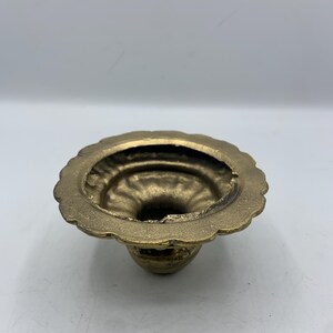 May include: A small, antique brass candle holder with a scalloped rim. The candle holder has a round base and a flared opening. The brass has a warm, golden hue and shows signs of age.