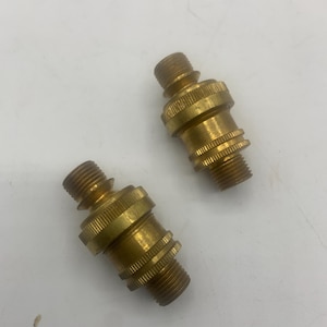May include: Two brass-colored threaded connectors. Each connector has a male thread on both ends and a ridged, cylindrical body. The connectors are likely used for plumbing or mechanical applications.