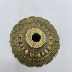 May include: A brass-colored, decorative lamp base with a floral design. The base has a round, fluted edge and a central, cylindrical socket. The finish has a slightly aged appearance.
