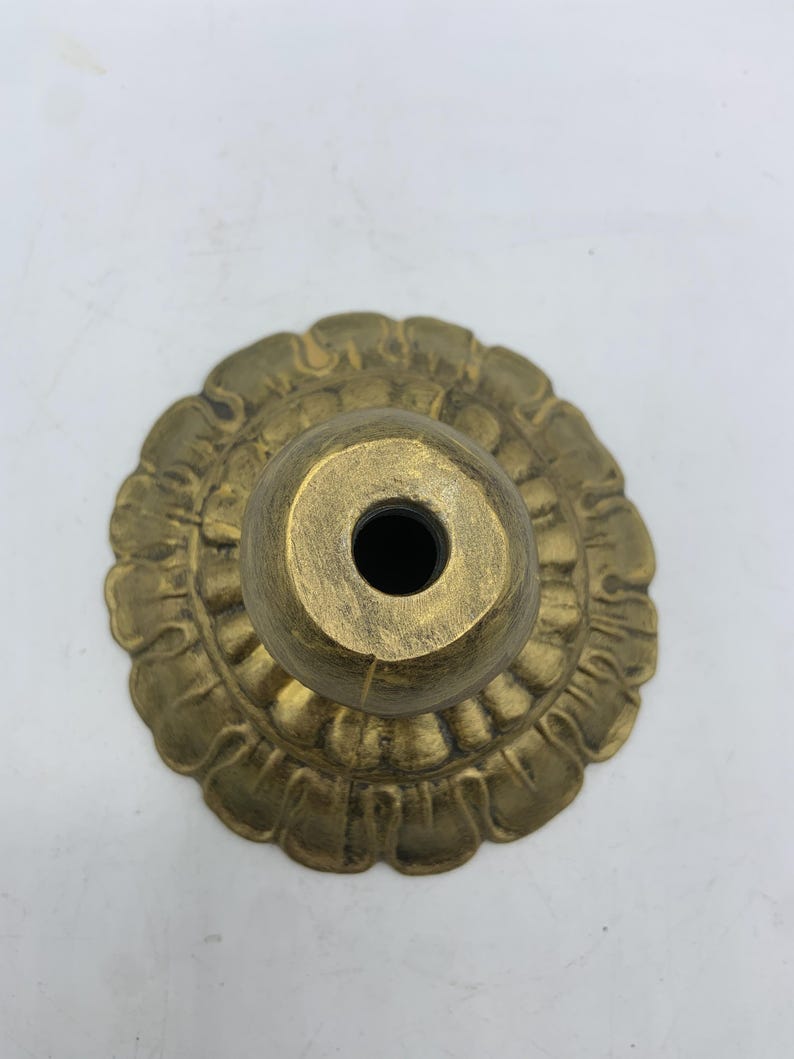 May include: A brass-colored, decorative lamp base with a floral design. The base has a round, fluted edge and a central, cylindrical socket. The finish has a slightly aged appearance.