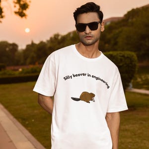 Könnte beinhalten: Ein weißes T-Shirt mit kurzen Ärmeln und einer Grafik eines braunen Bibers, der einen kleinen silbernen Schraubenschlüssel hält. Der Text "Silly beaver in engineering" ist in einer schwarzen, geschwungenen Schrift über dem Biber abgedruckt.