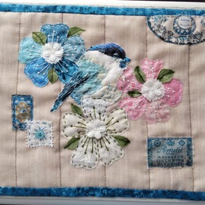 May include: A quilted textile with a blue bird and floral applique. The bird is blue and white, surrounded by blue, pink, and white flowers. The piece has a blue border and decorative patches.