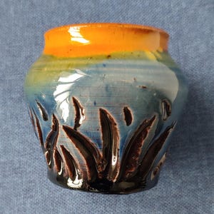 May include: A small, handmade ceramic vase with a vibrant colour scheme. The top rim is a bright orange, transitioning to blue and brown. The vase features a textured, carved design around the base.