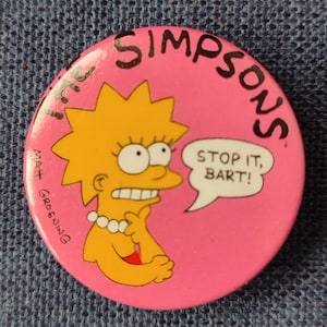 May include: A pink button featuring a yellow cartoon character with blonde hair and a speech bubble that reads "Stop it, Bart!" The button also says "The Simpsons" and "Matt Groening".