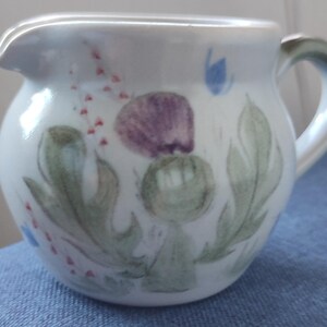 May include: A white ceramic milk jug with a green, purple and blue floral design. The design features a stylised thistle flower.