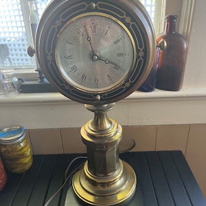 May include: An antique brass clock lamp with a round clock face featuring Roman numerals and a decorative black and gold border. The lamp has a detailed base and stem, and a black rectangular base.