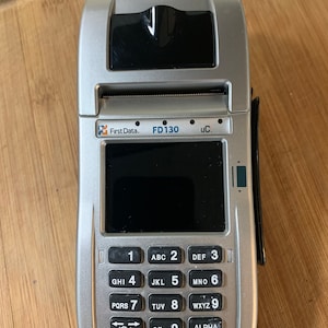 May include: A silver and black credit card processing terminal with a small screen and a keypad. The terminal has the text "First Data. FD130 uC." on the front.