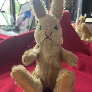 May include: A vintage, tan-colored plush bunny rabbit toy with black eyes and a pink nose. The rabbit is sitting on a red surface.