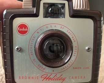 May include: A vintage Kodak Brownie Holiday camera with a brown plastic body and a silver metal front. The camera has a black lens with a silver ring around it. The text "Kodak", "Brownie Holiday", "DAKON LENS", and "MADE IN ROCHESTER, N.Y., U.S.A. BY EASTMAN KODAK COMPANY" is printed on the front of the camera.