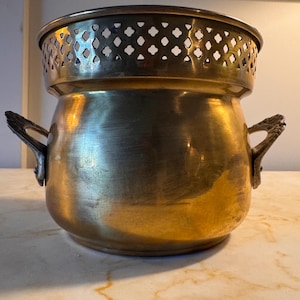 May include: A vintage brass pot with a decorative, pierced rim and two ornate handles. The pot has a rounded body and a warm, golden hue, suggesting age and use. The design includes a removable top with a lattice pattern.