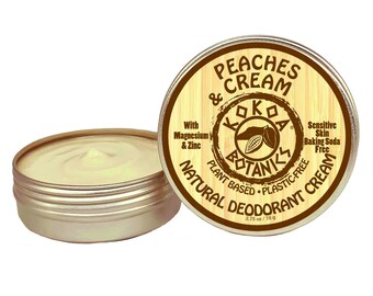 May include: A round tin of natural deodorant cream with a wooden lid. The lid has a brown label with the text "Peaches & Cream", "Koko Botanics", "Plant Based", "Plastic-Free", "Sensitive Skin Baking Soda Free", "Natural Deodorant Cream", and "2.75 oz/78 g".