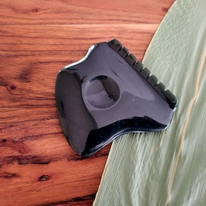May include: A black gua sha tool with a smooth, curved design. The tool has a circular indentation and a ridged edge. It rests on a wooden surface with a green leaf.