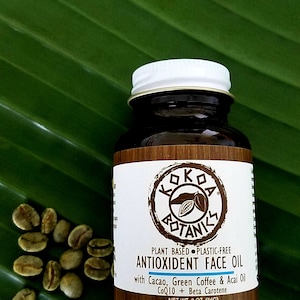 Op de afbeelding: Een bruine glazen fles met een wit deksel dat een plantaardige, plasticvrije antioxidant gezichtsolie bevat. Het etiket heeft een bruin logo met de tekst "Koko Botanics" en de woorden "Plant Based Plastic-Free Antioxidant Face Oil" met Cacao, Green Coffee & Acai Oil CoQ10 + Beta Carotene. De fles staat op een groen blad.