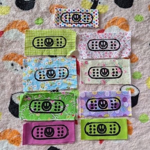 May include: Ten colorful fabric patches with a smiling face design. Each patch features a different background pattern and a black and white smiling face on a white bandage.