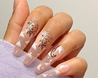 May include: Long, clear, fake nails with a white cloudy design and gold glitter accents. Some nails feature a black sun or moon design.