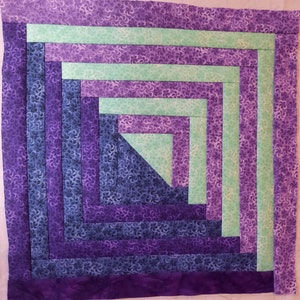May include: A quilt block with a log cabin pattern in shades of purple, blue, and green. The block is made up of strips of fabric sewn together in a concentric pattern.