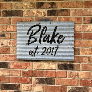 May include: A rectangular, corrugated metal sign with the name "Blake" in black script and "est. 2017" in a smaller, block font. The sign is mounted on a brick wall with varying shades of red, brown, and tan bricks.