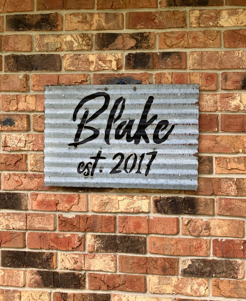 May include: A rectangular, corrugated metal sign with the name "Blake" in black script and "est. 2017" in a smaller, block font. The sign is mounted on a brick wall with varying shades of red, brown, and tan bricks.