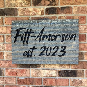 May include: A rectangular metal sign with the text "Fitt-Amerson est. 2023" in black lettering. The sign is made of corrugated metal and is mounted on a brick wall.