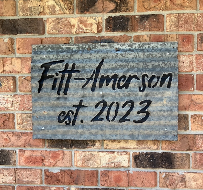 May include: A rectangular metal sign with the text "Fitt-Amerson est. 2023" in black lettering. The sign is made of corrugated metal and is mounted on a brick wall.