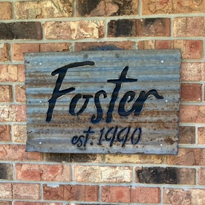 May include: A weathered metal sign with the name "Foster" cut out in a cursive font. Below the name is "est. 1990" in a smaller, matching font. The sign is mounted on a brick wall.