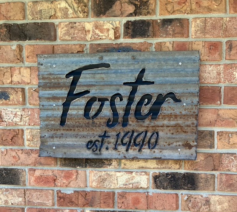 May include: A weathered metal sign with the name "Foster" cut out in a cursive font. Below the name is "est. 1990" in a smaller, matching font. The sign is mounted on a brick wall.