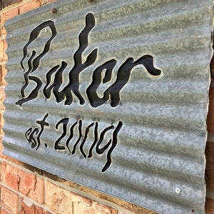May include: A galvanized metal sign with the name "Baker" cut out in a stylized font, with "est. 2004" below. The sign is mounted on a brick wall and has a corrugated texture.