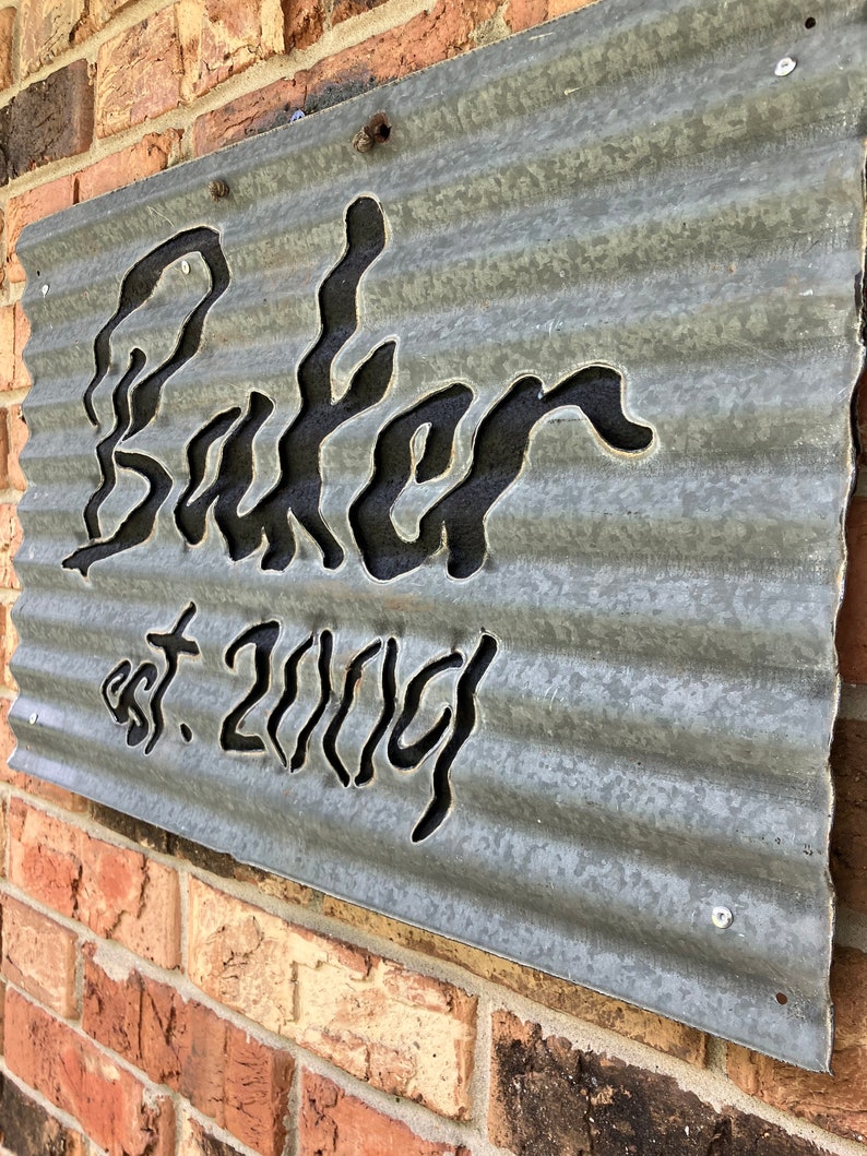 May include: A galvanized metal sign with the name "Baker" cut out in a stylized font, with "est. 2004" below. The sign is mounted on a brick wall and has a corrugated texture.