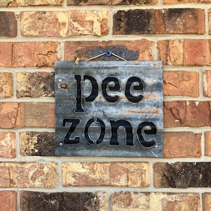 May include: A weathered metal sign with the words "Pee Zone" cut out in bold, black letters. The sign is attached to a brick wall with a small wire and a blue screw.