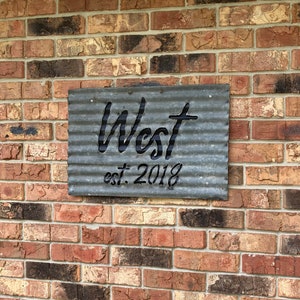 May include: A corrugated metal sign with the name "West" cut out, and the text "est. 2018" below. The sign is mounted on a brick wall with varying shades of red, brown, and gray bricks.