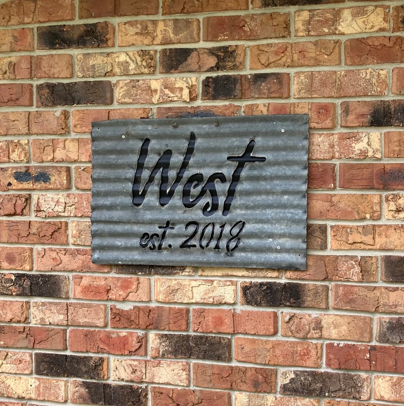 May include: A corrugated metal sign with the name "West" cut out, and the text "est. 2018" below. The sign is mounted on a brick wall with varying shades of red, brown, and gray bricks.