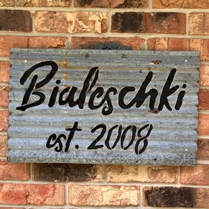 May include: A galvanized metal sign with the name "Bialeschki" in black script and the text "est. 2008". The sign is mounted on a brick wall with varying shades of red, brown, and tan bricks.