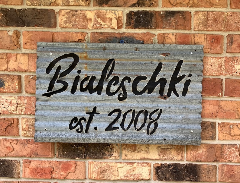 May include: A galvanized metal sign with the name "Bialeschki" in black script and the text "est. 2008". The sign is mounted on a brick wall with varying shades of red, brown, and tan bricks.