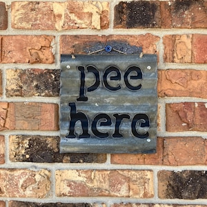May include: A rustic metal sign with the words "pee here" in black lettering on a corrugated metal background. The sign is hanging on a brick wall.