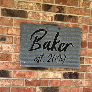May include: A rectangular metal sign with the name "Baker" in a cursive font and the text "est. 2009" in a smaller, block font. The sign is mounted on a brick wall. The metal has a corrugated texture.