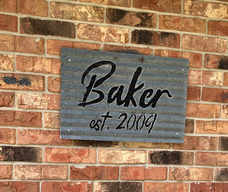 May include: A rectangular metal sign with the name "Baker" in a cursive font and the text "est. 2009" in a smaller, block font. The sign is mounted on a brick wall. The metal has a corrugated texture.