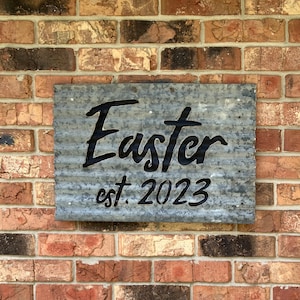 May include: A galvanized metal sign with the word "Easter" in black script and the text "est. 2023". The sign is mounted on a brick wall. The sign has a corrugated texture.