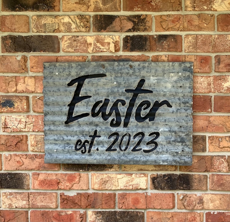 May include: A galvanized metal sign with the word "Easter" in black script and the text "est. 2023". The sign is mounted on a brick wall. The sign has a corrugated texture.