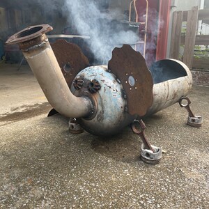 May include: A metal elephant sculpture with a rusty patina. The sculpture is made from repurposed metal parts, including a large pipe for the trunk and engine parts for the legs. The sculpture is sitting on a concrete surface.
