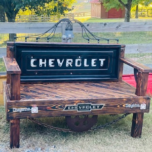 May include: A rustic wooden bench with a black metal Chevrolet tailgate as the backrest. The bench has a wooden seat with a distressed wood finish and a metal chain connecting the two sides. The bench has a Chevrolet logo on the front and the word "Cheyenne" on each side.